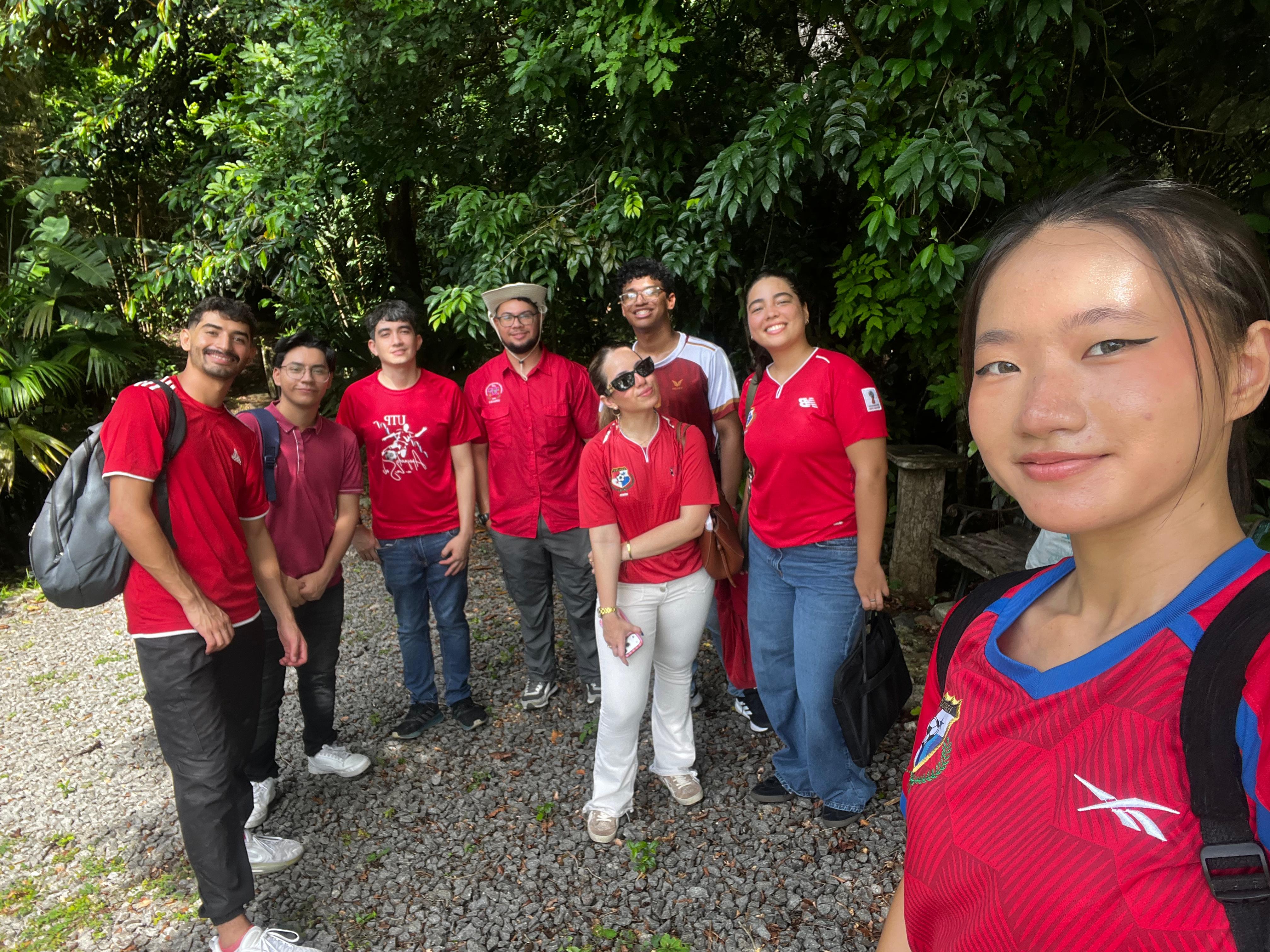 Jacqueline Liu (Panabuddy) a la derecha toma una selfi junto a un grupo de siete compañeros sonrientes, celebrando a la sele, mientras caminan por un sendero de piedra. El grupo está rodeado de una densa y verde vegetación selvática que enmarca la escena con un ambiente natural y alegre.
