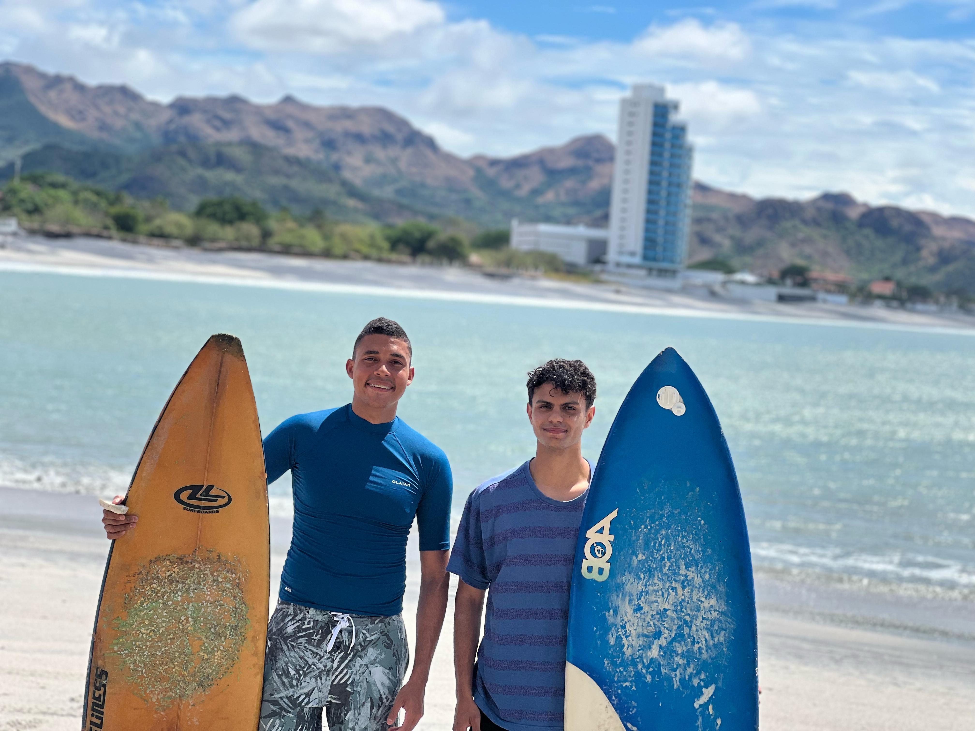 Gregory Lopez (a la izquierda) y Paolo Galvez (a la derecha) en una playa de arena clara sosteniendo sus respectivas tablas de surf, una amarilla y otra azul. El fondo formal y profesional muestra una bahía de aguas turquesas rodeada de montañas y un moderno edificio de gran altura bajo un cielo parcialmente nublado.