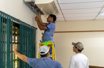 Tres estudiantes realizan mantenimiento a una unidad de aire acondicionado montada en la pared de un aula